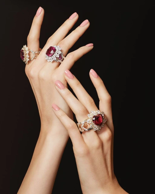Close-up of a woman's hands adorned with three large ruby and diamond rings, presented against a dark background.
