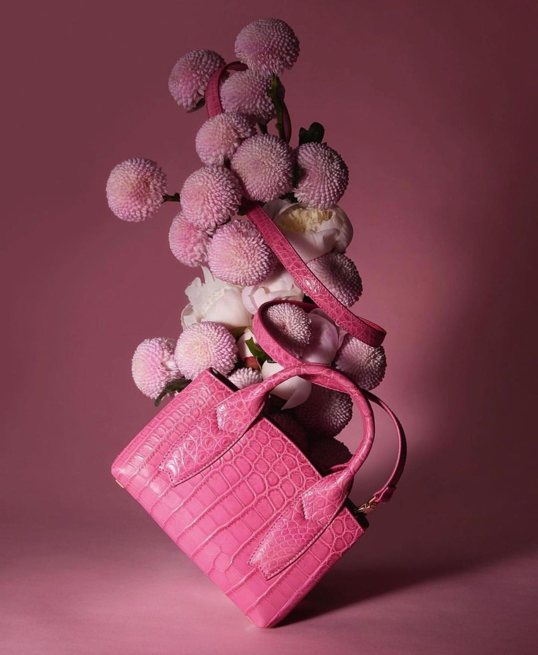 Fashion shot of pink crocodile-texture purse on pink background with pink pompom flowers. Bright, vibrant, and modern.