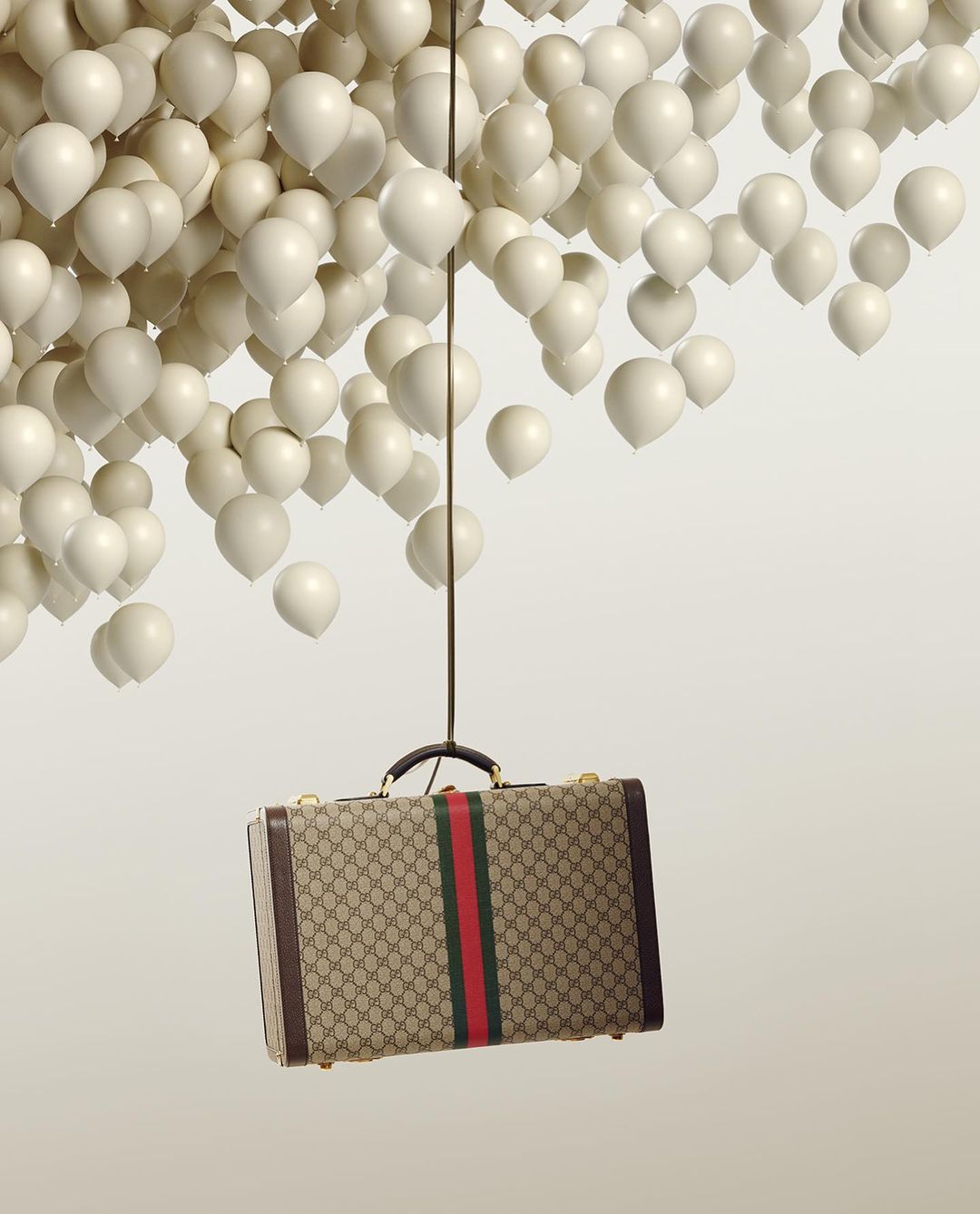A tan Gucci suitcase is suspended by a thin black rope attached to a cluster of white balloons on a neutral background in a studio shot.