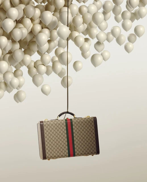 A tan Gucci suitcase is suspended by a thin black rope attached to a cluster of white balloons on a neutral background in a studio shot.