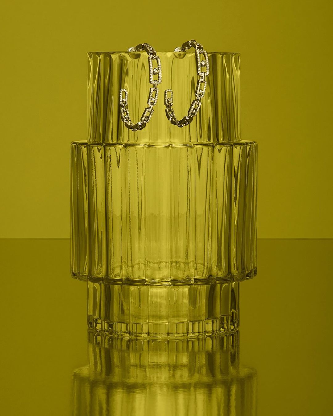 Product still life of a pair of silver chain-link hoop earrings displayed on a ridged clear vase against a solid yellow background.