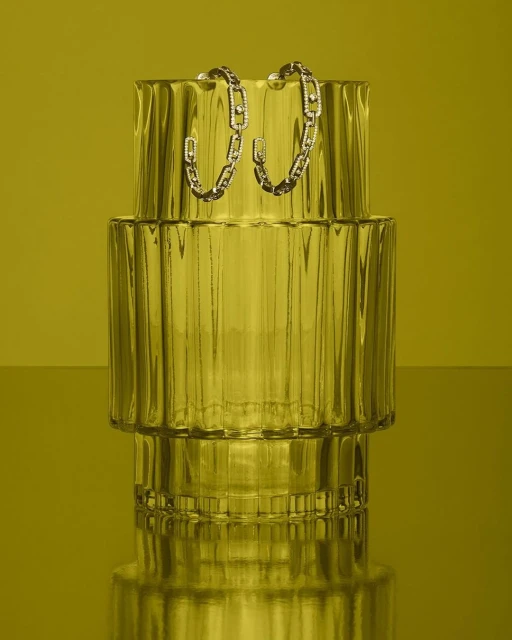 Product still life of a pair of silver chain-link hoop earrings displayed on a ridged clear vase against a solid yellow background.