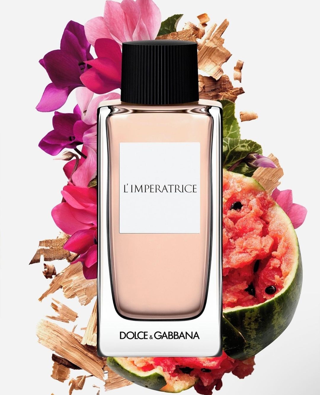 Dolce & Gabbana L'Imperatrice perfume bottle showcased with pink flowers, wood splinters and watermelon slices against a light background.