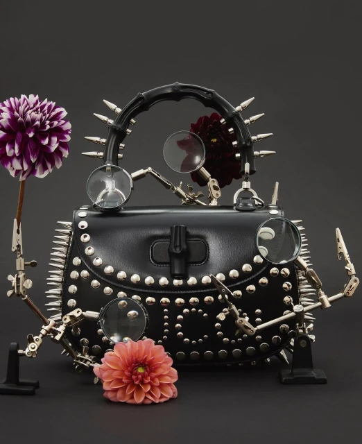 Studded black leather purse surrounded by flowers and articulated magnifying glass arms on dark background, product photography.