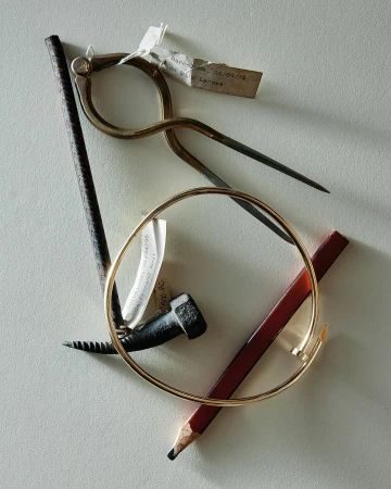 Still life of drafting compass, bracelet, nail, pencil and metal rod on neutral surface with label and geometric composition.