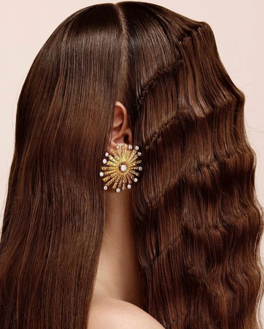 Close-up of a woman with long brown hair styled with a braid, wearing a large, sunburst-style gold earring with pearl and diamond accents.