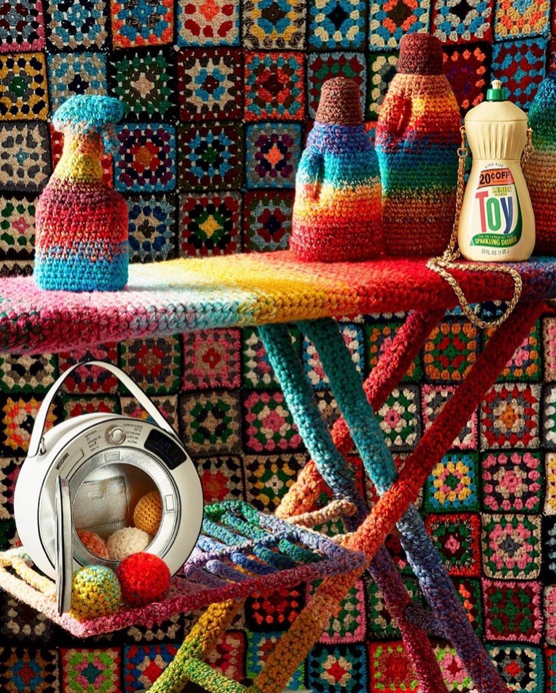 Colorful crochet craftwork featuring household objects, a mini washing machine and a blanket-style backdrop, all handcrafted with vibrant yarn.