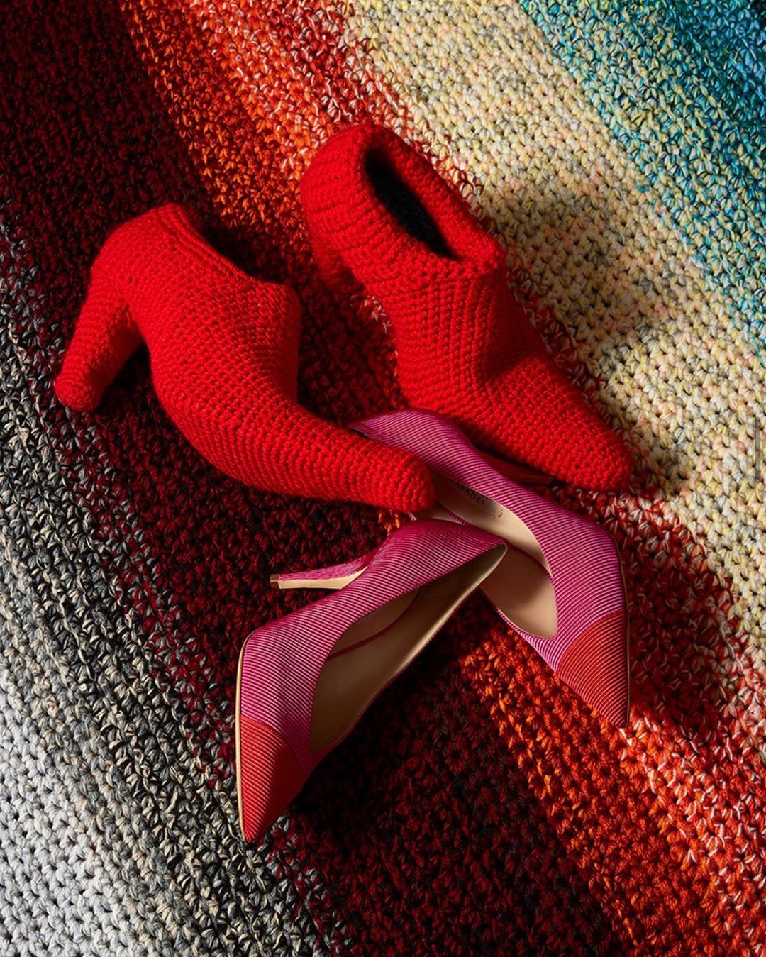 Still life of a pair of pink stiletto heels with cap toes, resting next to a pair of red slipper heels on a striped textile.