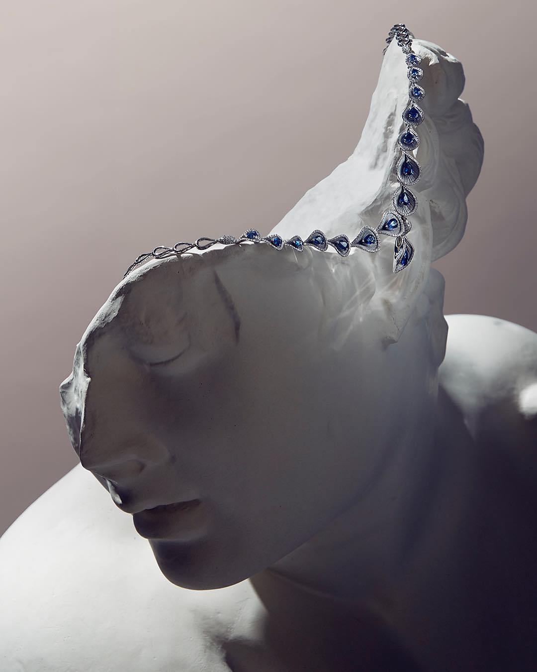 Close-up of a white sculpture wearing a blue teardrop necklace against a grey background, showcasing jewelry and classical art.
