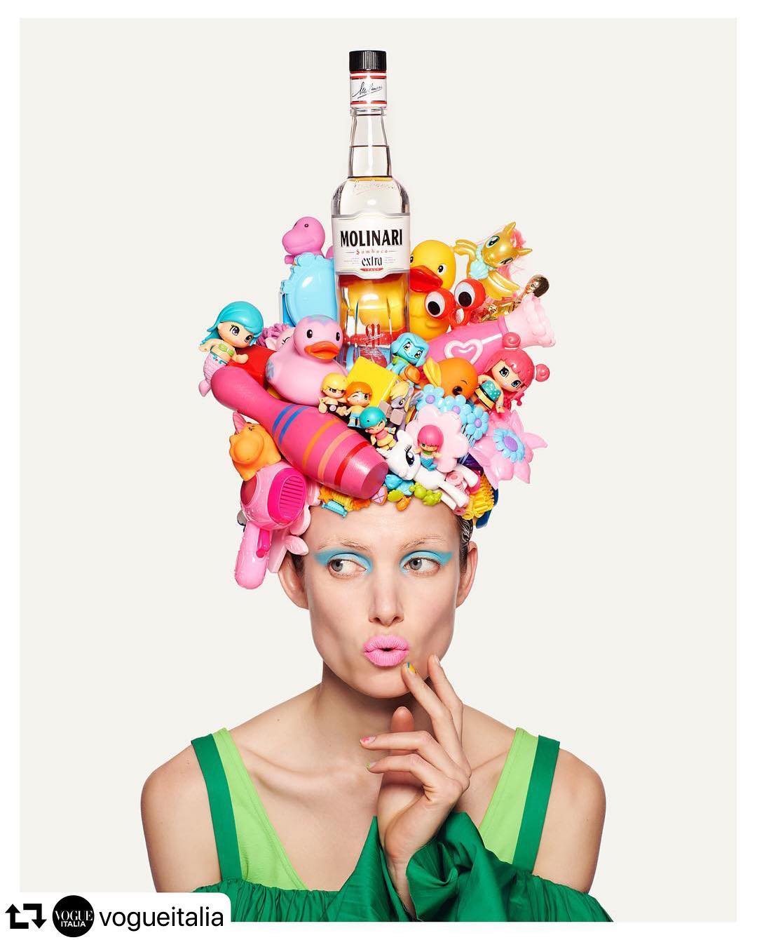 Adult woman with quirky pastel makeup wears an elaborate hat of childrens toys and clear bottle, against a white backdrop.