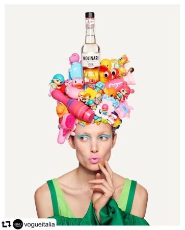Adult woman with quirky pastel makeup wears an elaborate hat of childrens toys and clear bottle, against a white backdrop.