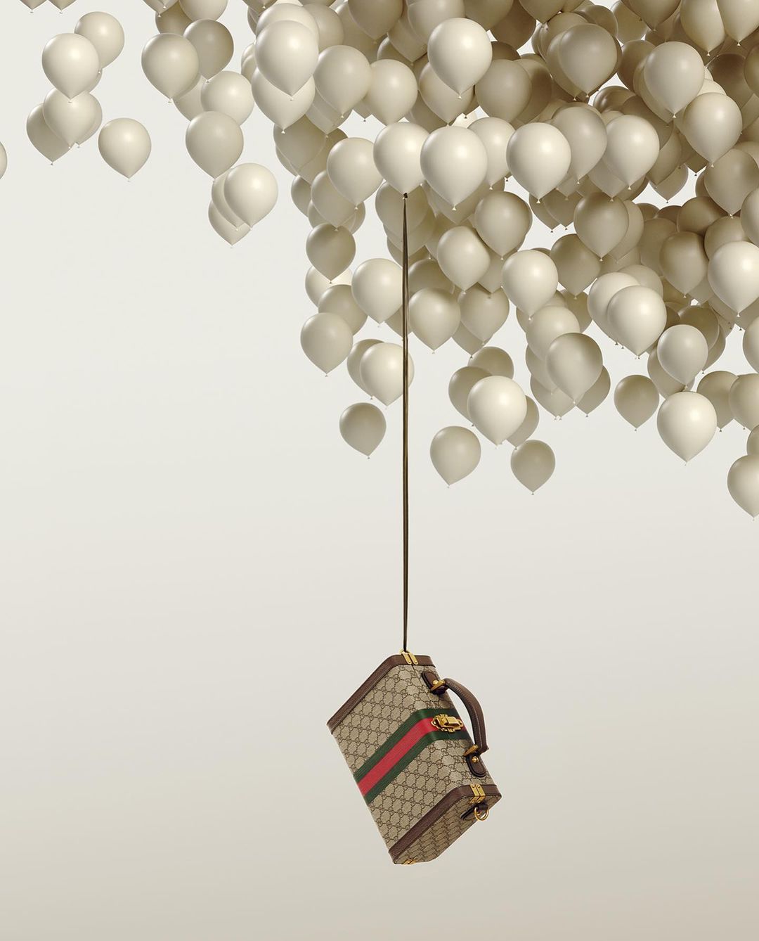Gucci suitcase suspended by strings tied to numerous white balloons against a light gradient backdrop in a studio setting.