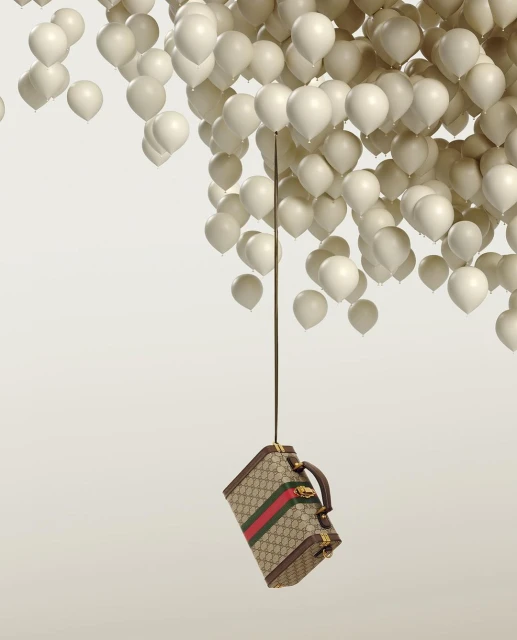 Gucci suitcase suspended by strings tied to numerous white balloons against a light gradient backdrop in a studio setting.