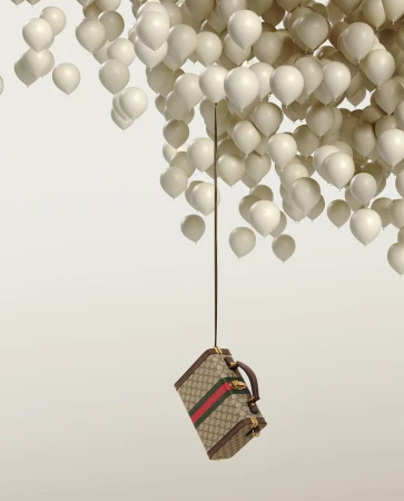 Gucci suitcase suspended by strings tied to numerous white balloons against a light gradient backdrop in a studio setting.