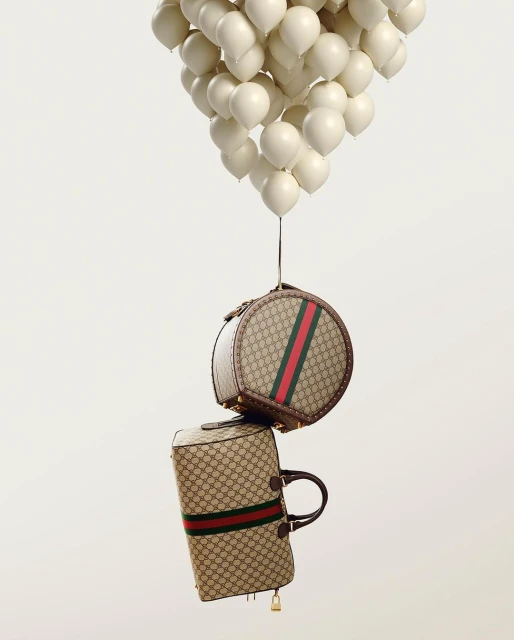 Gucci luggage floats with a bundle of white balloons against a white background in a fashion product still life shot.