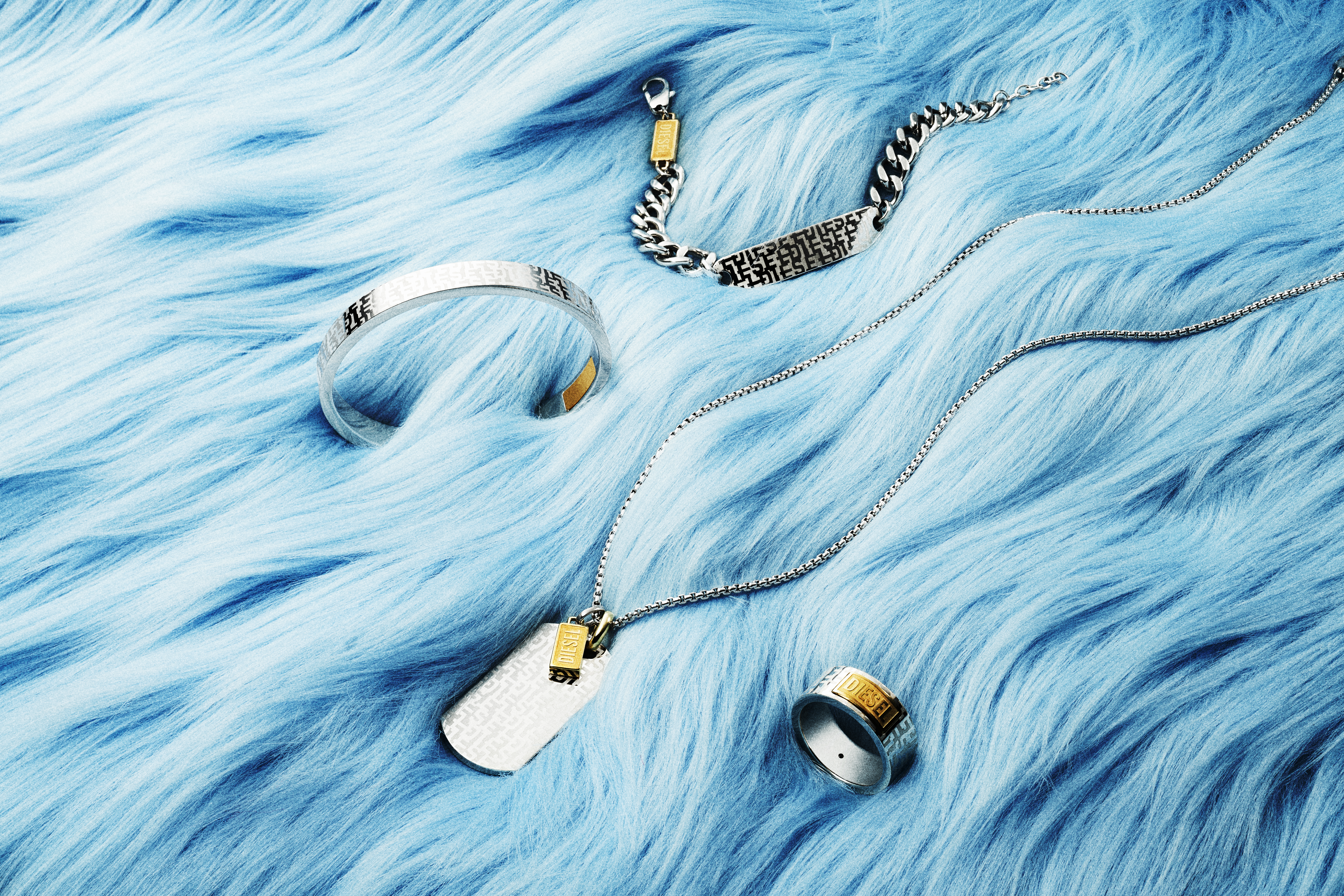 Silver-and-gold toned jewelry on a light blue faux-fur background, including a ring, bracelet, dog tag pendant, and chain.