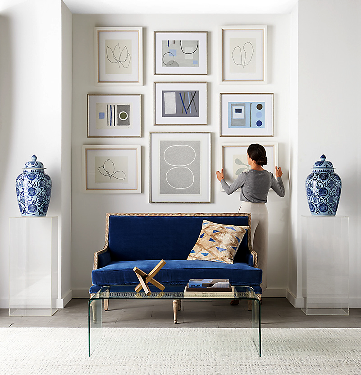 Interior of a living room with blue settee under framed art, flanked by decorative vases and set on a white rug and glass coffee table.