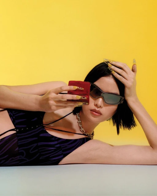 Woman lying down in swimsuit with sunglasses holding smartphone. Yellow backdrop with soft lighting, fashion and beauty portrait.