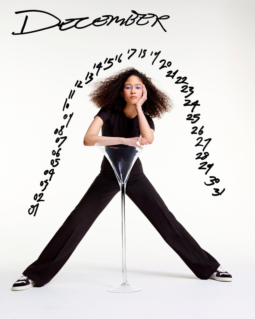 A young adult female wearing black pants and a black top poses in front of a white backdrop, with black writing above her. 