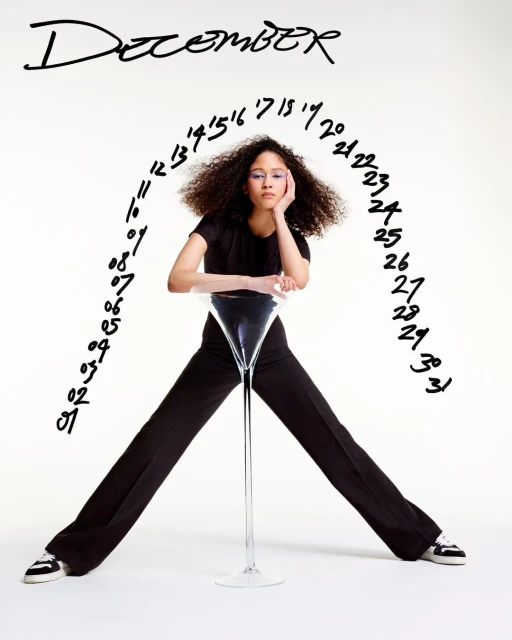 A young adult female wearing black pants and a black top poses in front of a white backdrop, with black writing above her. 