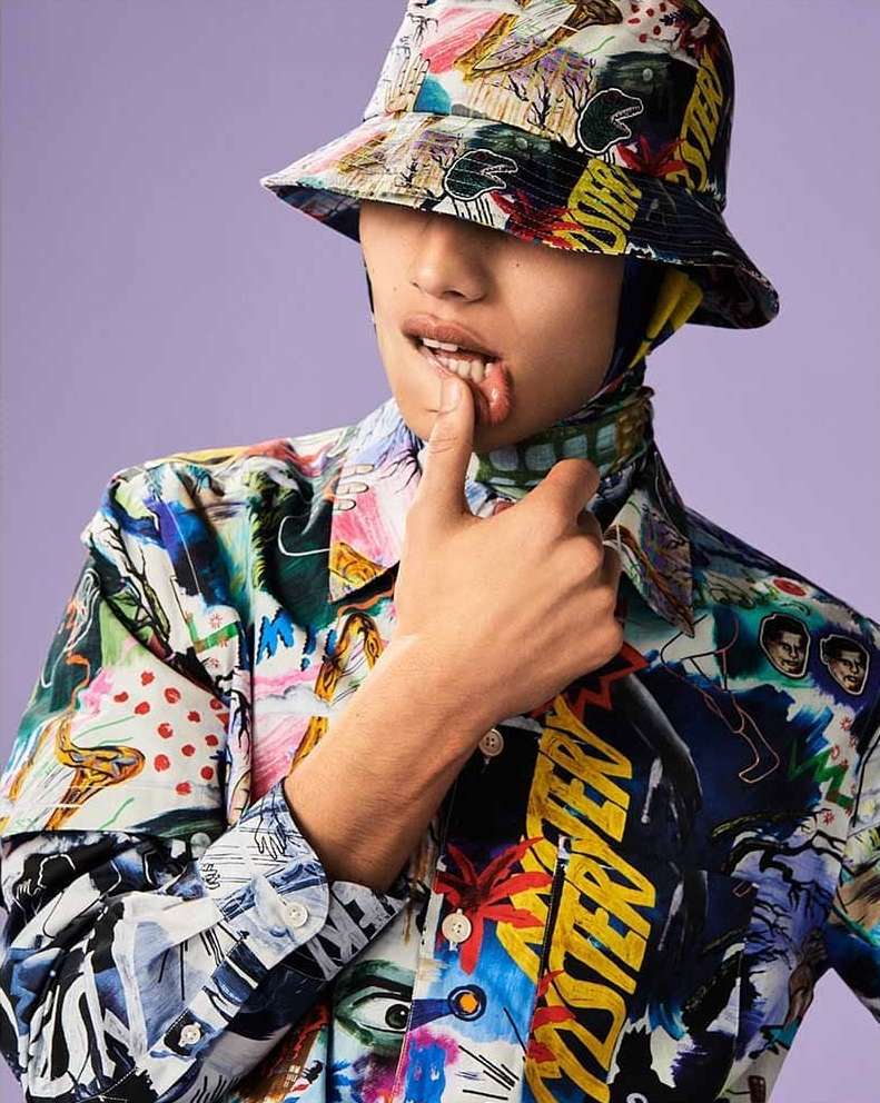 Person wearing a colorful patterned bucket hat and matching shirt looks at the camera with a finger near their mouth on a purple background.