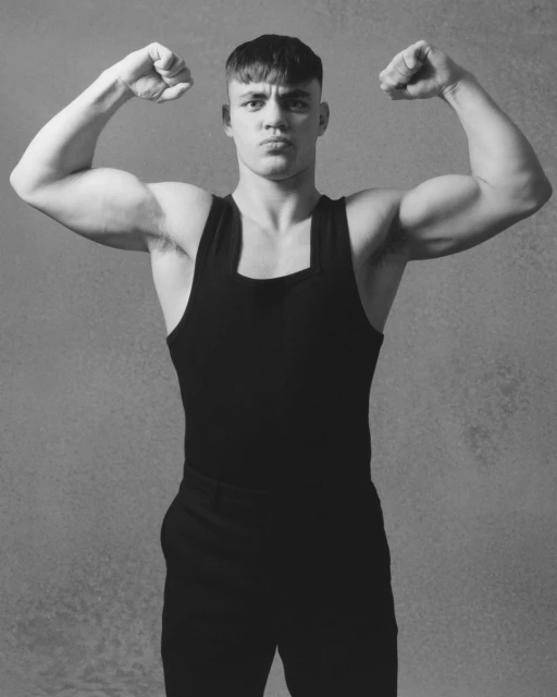 Muscular man flexing biceps in black tank top and pants against a textured gray background in a black and white studio portrait.