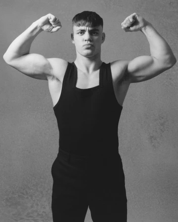 Muscular man flexing biceps in black tank top and pants against a textured gray background in a black and white studio portrait.