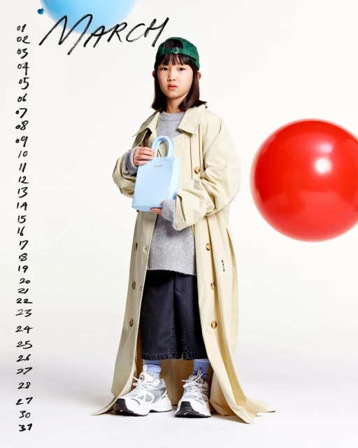 A young girl in a beige trench coat holds a light blue purse. The photo, taken in a studio, has a calendar and colorful balloons.