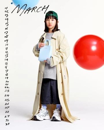 A young girl in a beige trench coat holds a light blue purse. The photo, taken in a studio, has a calendar and colorful balloons.