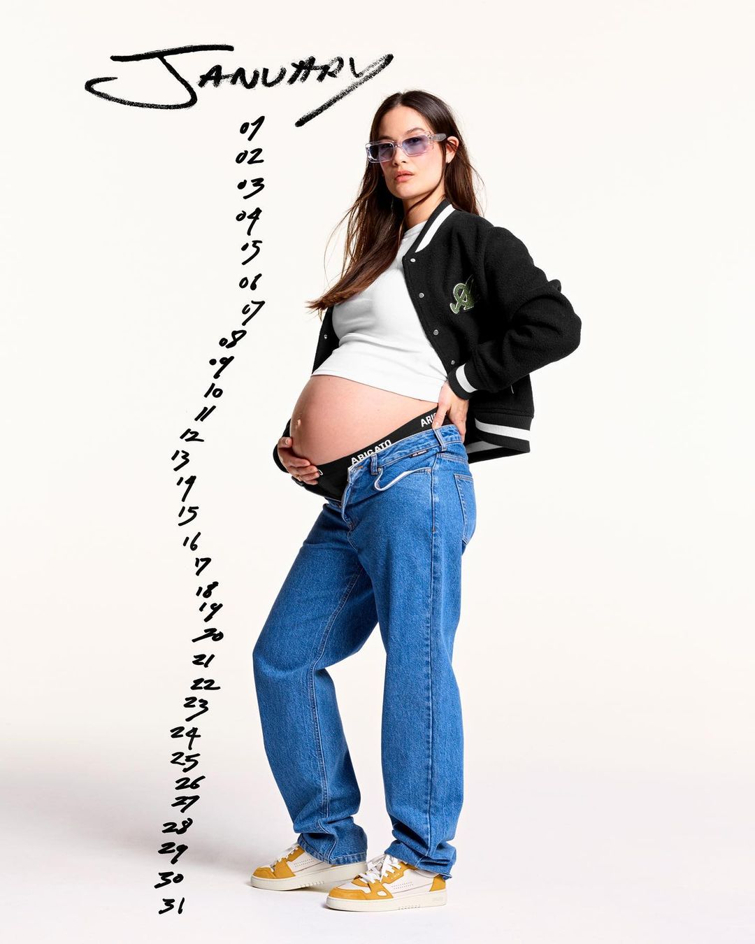 A pregnant adult female in a casual outfit poses in a photo studio against a white backdrop with handwritten text in the foreground.