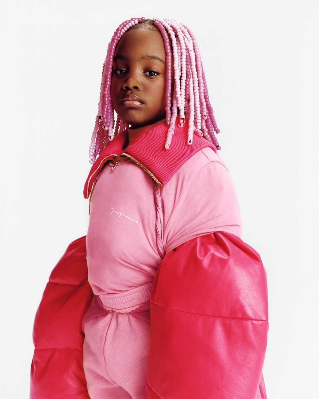 Child in a pink puffer jacket with oversized sleeves and braided hair with beads posing in a photo studio against a white backdrop.