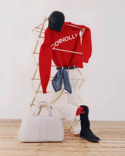 Stylized flat lay of fashion items including a red Connolly sweater, white pants, black hat and shoes, and a white duffel bag.