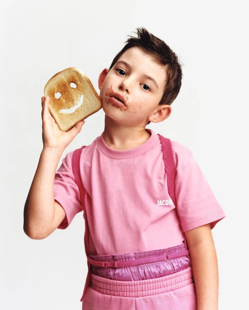 Young child in a pink outfit holds toast with a smiley face near his cheek, with chocolate on his face, against a white backdrop.