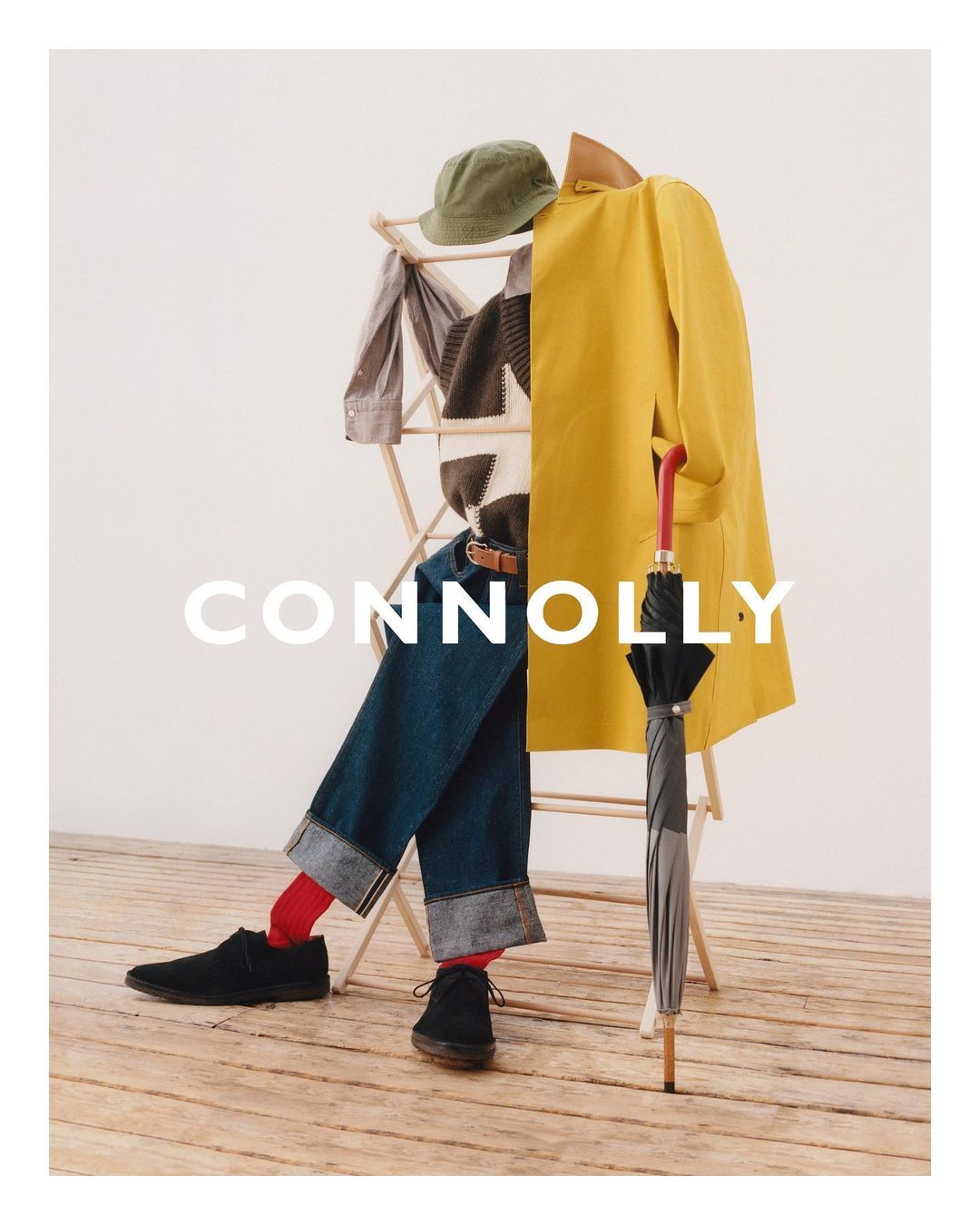 Still life of men's clothing including a yellow raincoat, blue denim trousers, a tweed jacket and an umbrella on a wooden floor.