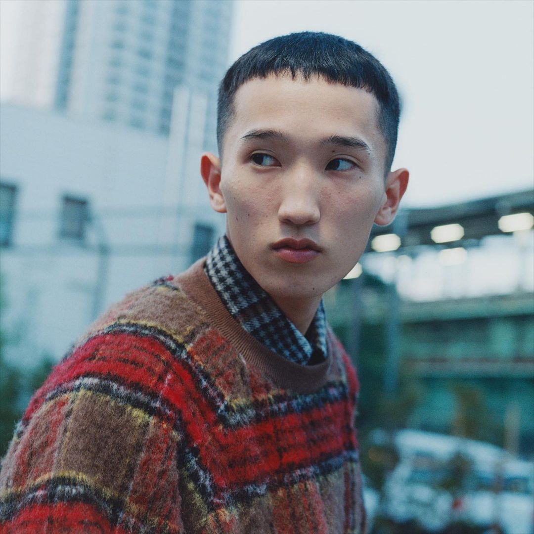 Portrait of an Asian man with short hair wearing a patterned sweater. Urban setting with buildings and a bridge in the background.