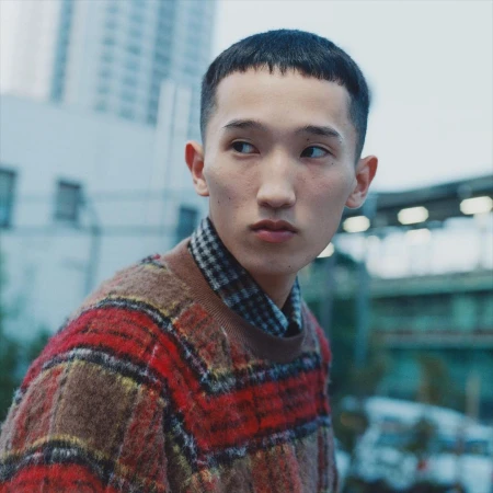Portrait of an Asian man with short hair wearing a patterned sweater. Urban setting with buildings and a bridge in the background.