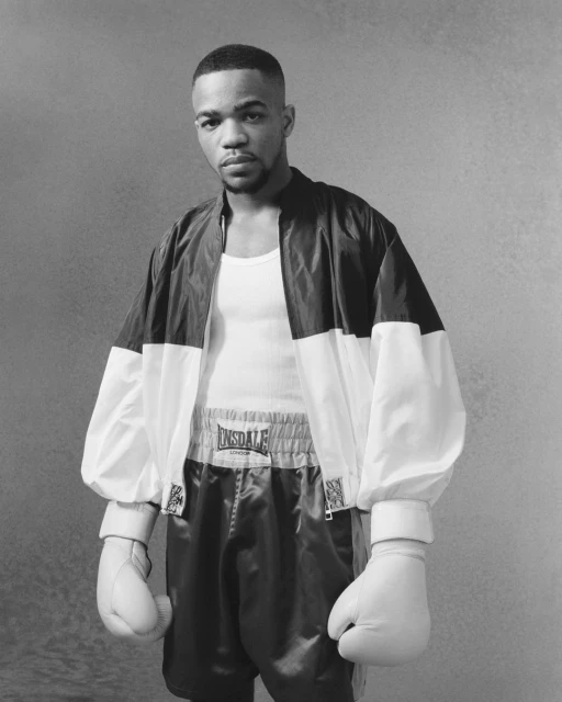 Black and white portrait of a boxer in boxing shorts and gloves, wearing a jacket, standing in front of a grey background.