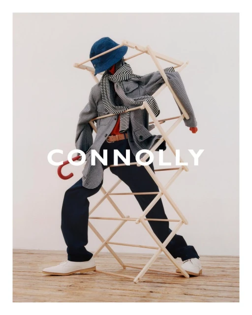 Fashionable individual in a gray jacket, blue hat, and dark pants poses with a wooden clothes drying rack indoors.