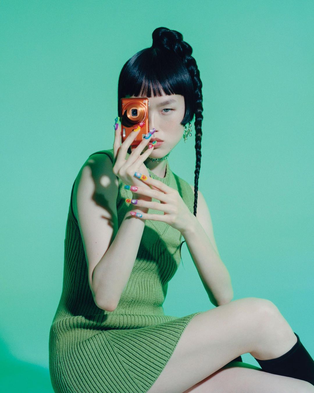 Asian woman with stylish braided hair poses with an orange compact camera covering one eye, wearing a green dress against a green backdrop.