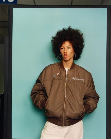 A person with voluminous dark curly hair wearing a brown bomber jacket posing against a pale blue backdrop.