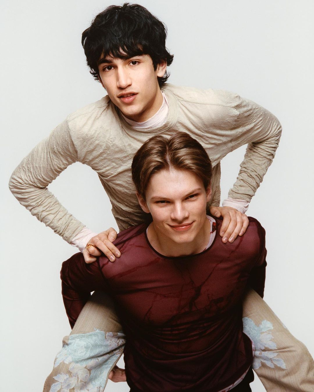 Two young adult men, one giving a piggyback ride, both smiling, dressed in casual attire, set against a plain white backdrop. 
