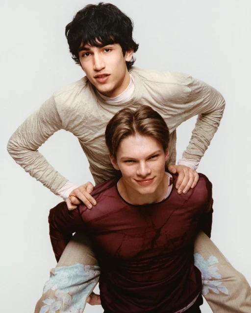 Two young adult men, one giving a piggyback ride, both smiling, dressed in casual attire, set against a plain white backdrop. 