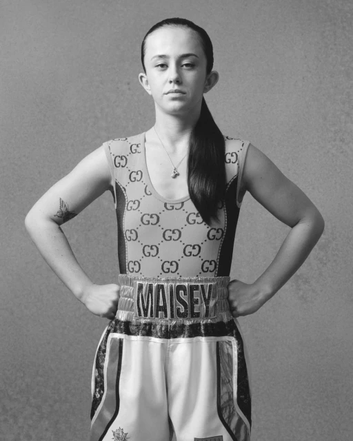 A young adult woman poses in a boxing outfit with a Gucci-patterned top and boxing shorts, standing against a plain gray backdrop in a studio shot.
