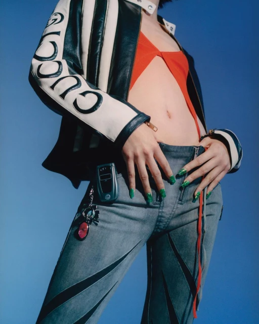 Cropped shot of a slender adult female wearing a Gucci jacket, red top, jeans, and green nails, posing against a blue background.