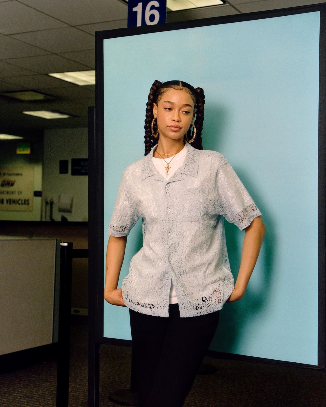 Portrait of a young adult female with braided pigtails posing in a light blue button-down shirt and black skirt in an office setting.