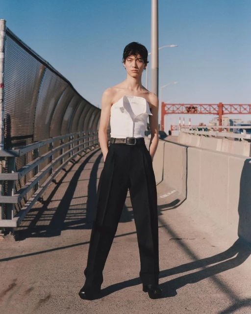 Fashion shot of a person in a strapless top and black pants standing on a bridge on a sunny day with a city skyline in the background.
