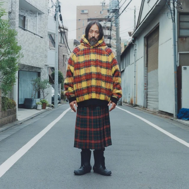 Adult male with a beard poses on an empty street wearing a red and yellow plaid hoodie and green plaid kilt with buildings in the background.