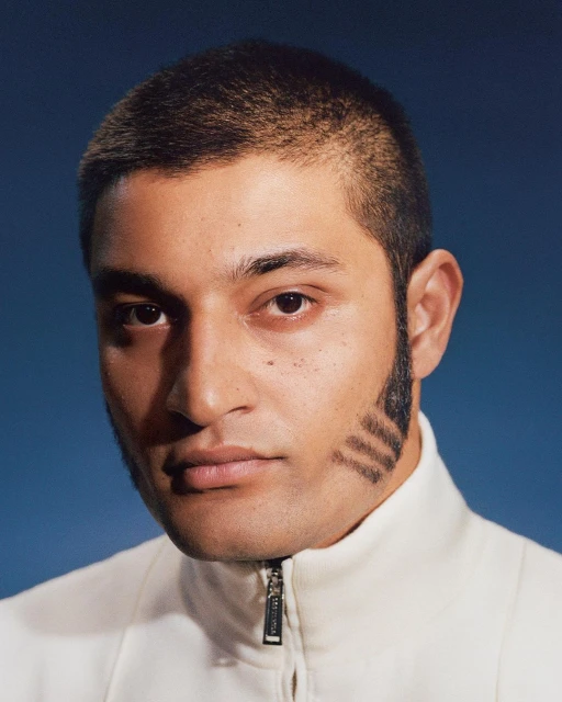 Close-up portrait of a man with short hair and distinctive sideburns wearing a white jacket against a blue background. Fashion-forward.