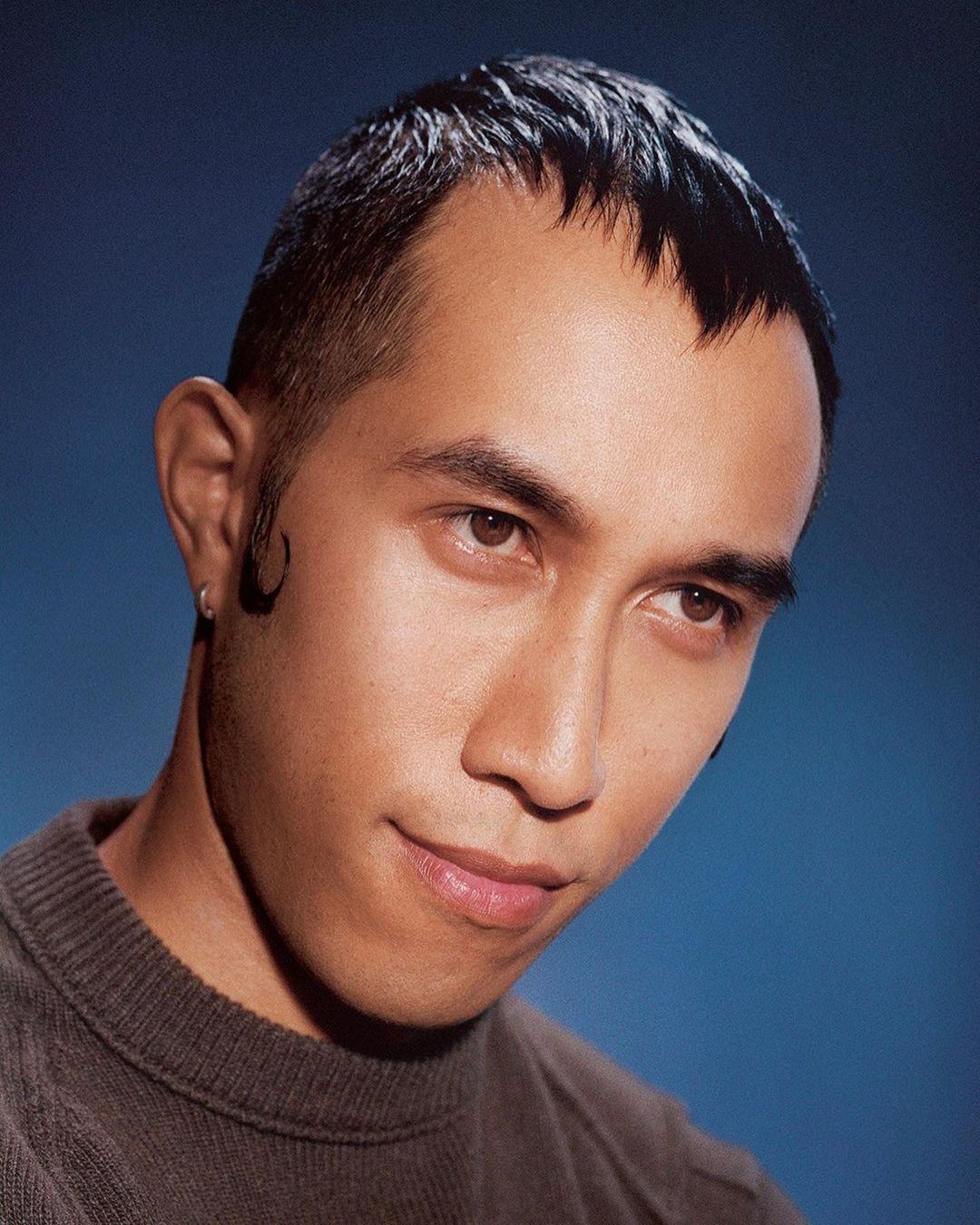 Portrait of an Asian man with short dark hair and a small earring against a blue background