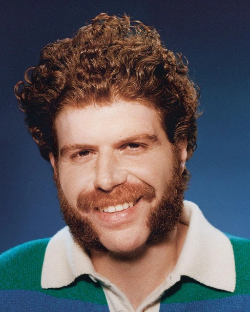 Close-up studio portrait of a man with curly red hair and a full beard, wearing a striped rugby shirt against a gradient blue background.